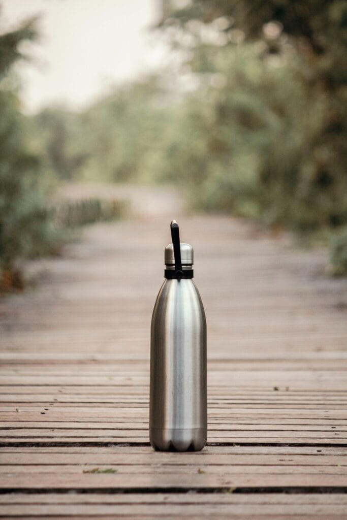 A stainless steel water bottle on a wooden path in an outdoor setting, surrounded by greenery.